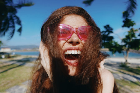 Woman smiling joyfully outdoors with sunglasses and long hair, set against a blue sky and greenery
