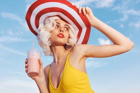 Young woman in a bright yellow swimsuit and a large red and white sun hat, holding a pink drink,...