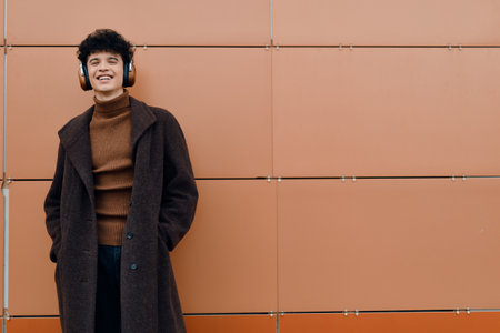 Fashion portrait of a young man wearing headphones, standing in front of an orange wall, smiling at the cameraの写真素材