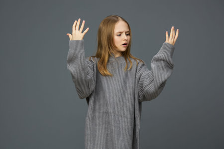 Young woman in oversized gray sweater expressing frustration with raised hands against a minimalist gray background, showcasing deep emotions and contemporary fashionの写真素材