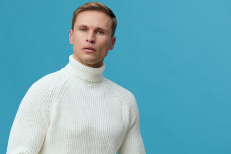 Portrait of a serious young man with short hair wearing a cozy white turtleneck sweater, looking thoughtfully at the camera against a plain blue background, studio shot, minimalistic aesthetic, fashion conceptの写真素材