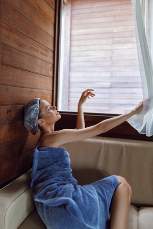 woman wrapped in blue towel and shower cap relaxing on beige sofa by wooden walls and soft sunlight through sheer curtains in cozy room interiorの写真素材