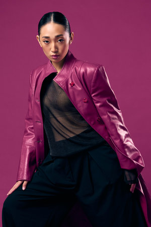 Fashion asian woman in purple leather jacket and black pants posing on purple background with eyes closed and hands on hipsの写真素材