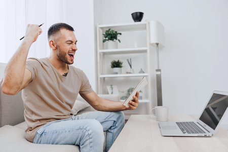Happy man celebrating success while using a tablet in a bright modern living room. Energetic mood, casual clothes, and minimalistic decor create a positive atmosphere.の写真素材