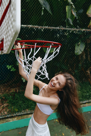 Joyful young woman playing basketball on an outdoor court, wearing a white outfit, showcasing excitement and a vibrant atmosphere amid lush green backgroundの写真素材