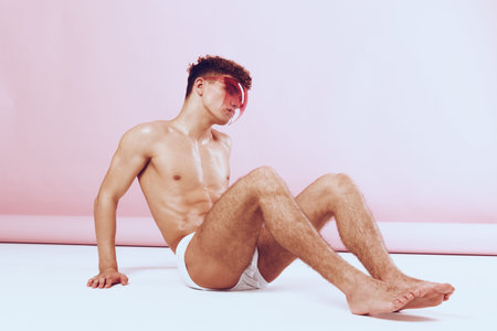 Young man with stylish sunglasses sitting against a pastel pink background, showcasing confidence...