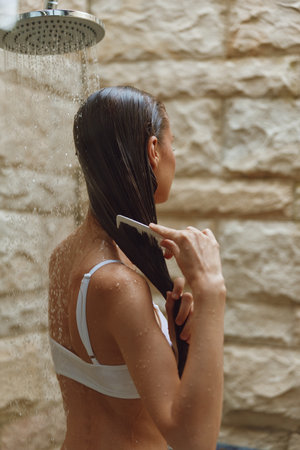 woman combing wet hair under outdoor shower with stone wall background, natural light, summer wellness moment, healthy lifestyle conceptの写真素材