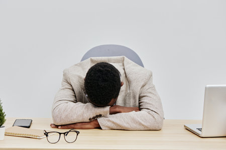 Businessman with dark hair hunched over desk resting head on arms exhaustion stress tiredness office...
