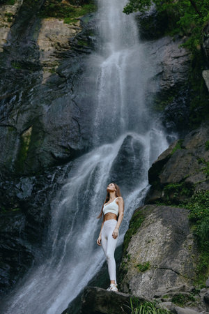 Woman standing near a large waterfall in white fitness wear surrounded by rock formations and lush greenery creating a serene outdoor atmosphere.の写真素材
