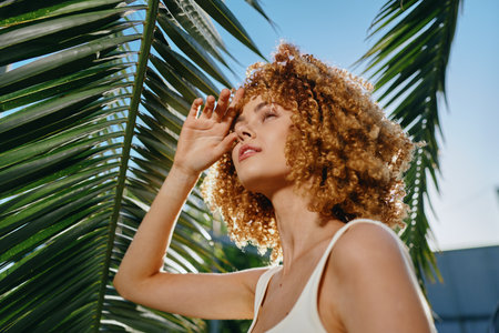 curly woman enjoying sunlight under palm leaves, with natural makeup and casual white top, capturing a bright and sunny summer mood outdoors.の写真素材