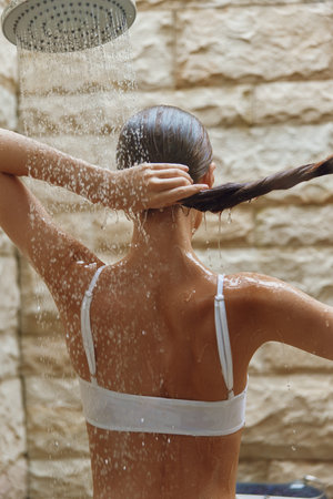 woman taking shower outdoors with flowing water, wet hair, stone wall background, natural light, refreshing moment, summer wellness.の写真素材