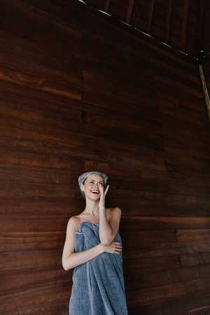 smiling woman wrapped in towel with headband stands against dark wooden wall, expressing joy and relaxation indoors in a cozy atmosphereの写真素材