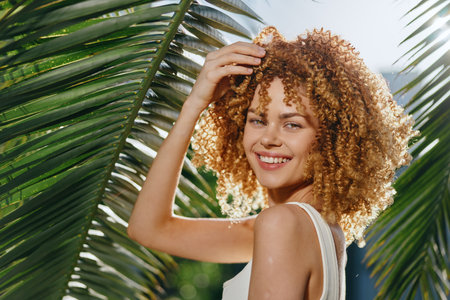 smiling woman with curly hair poses joyfully in sunlight amidst tropical palm leaves, wearing a white tank top and natural makeup, bright summer moodの写真素材