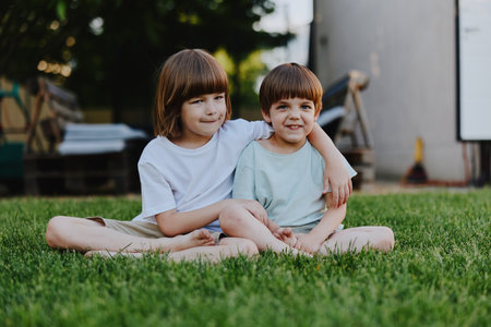 Two cheerful children sitting on green grass, enjoying a sunny day, displaying sibling affection and happiness, with a soft natural background.の写真素材
