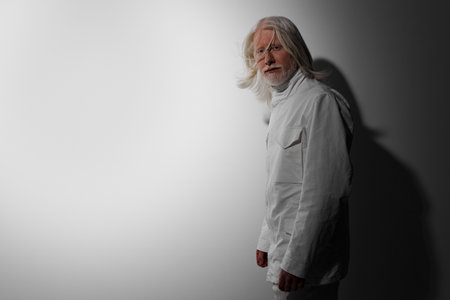 Portrait of an elderly man with long white hair, dressed in a white outfit against a light gray...