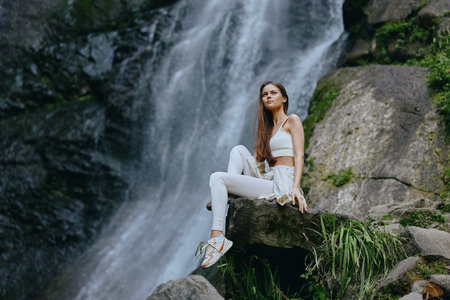 Young woman sitting on rock near waterfall in forest, wearing white activewear and sneakers, enjoying nature and outdoor adventure, peaceful and relaxed atmosphere.の写真素材