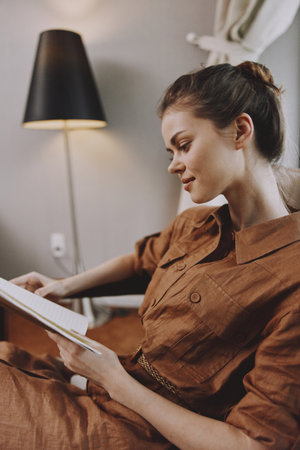 Young woman in a brown linen dress reading a book in a cozy living room, showcasing relaxation and warmth, ideal for lifestyle and home decor themesの写真素材