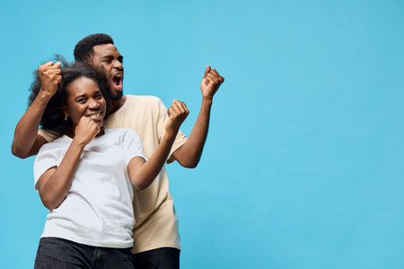 Happy diverse couple celebrating success with joyful expressions against a solid blue background, wearing casual outfitsの写真素材