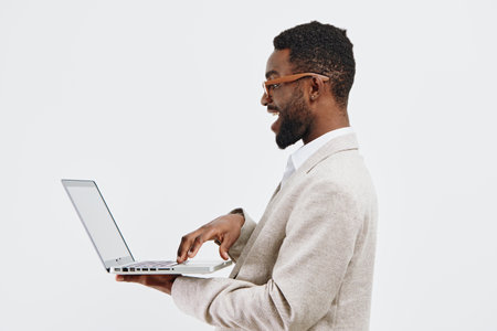 Smiling young African American man in business casual attire using laptop against plain white background. Studio shot, technology, communication, professional, desktop, modern lifestyle, confident.の写真素材