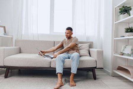 Focused man working on a laptop in a cozy living room, adorned with greenery and soft natural light, captures a modern, tranquil lifestyle.の写真素材