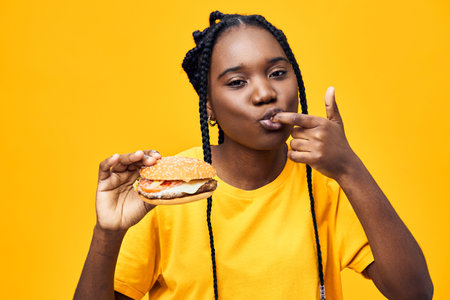 Happy young Black woman enjoying a delicious burger against a bright yellow background, showcasing...