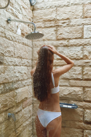 Woman showering outdoors under rainfall in a natural stone tiled shower area with relaxed posture and long wet hair, enjoying summer freshness and calm environment.の写真素材
