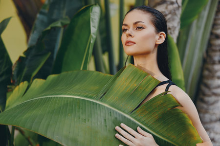 Exotic woman holding a large green leaf in front of her face against palm trees backgroundの写真素材