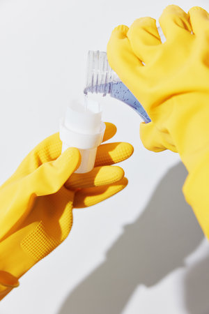 Woman in yellow gloves pouring cleaning solution from bottle into container on white surfaceの写真素材