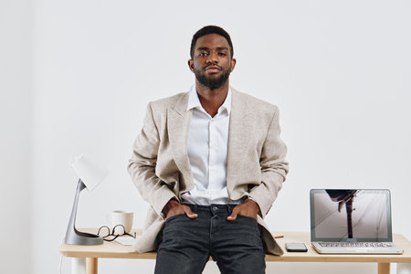 Confident young man with dark skin, stylish beige blazer, white shirt, and jeans sitting on desk with modern workspace setup, minimalistic background, and natural lighting. Business casual fashion, professional attitude, indoor portrait.の写真素材