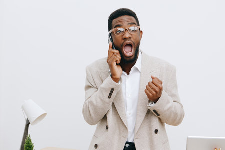 Professional young African American man in beige blazer and glasses shows surprise while talking on...