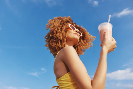 Young woman enjoying a refreshing drink under a bright blue skyの写真素材
