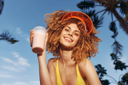 Smiling woman with curly hair holding a pink smoothie against a sunny sky with palm trees