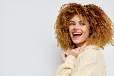 Smiling woman with curly hair showing happiness against a plain light background, wearing a cozy sweater, exuding joy and confidence in a portrait style settingの写真素材