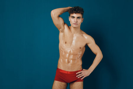 Confident athletic man posing in red swimwear against a teal background, showcasing toned physique and healthy lifestyle. Fitness and body positivity concept.の写真素材