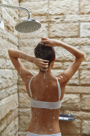 woman showering outdoors under rain showerhead with stone wall background, natural light, health and...