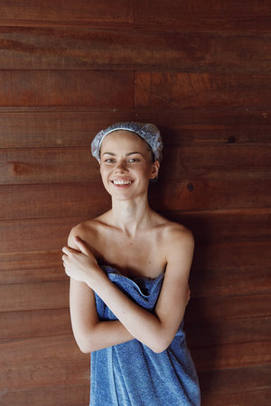 young woman wrapped in blue towel smiling happily with shower cap on wooden background, wellness, relaxation, spa, skincare routine, self-care conceptの写真素材
