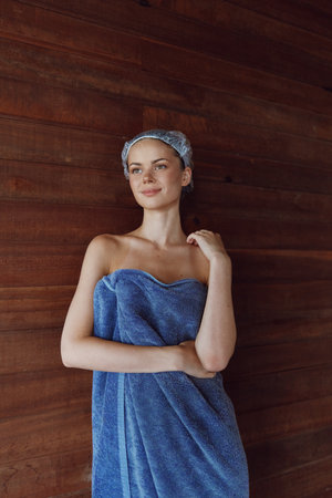 young woman wrapped in blue towel with shower cap standing against wooden wall, serene expression, natural light, wellness and spa atmosphereの写真素材