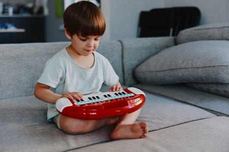 Young boy playing a red keyboard, sitting on a cozy sofa in a stylish living room, showcasing creativity and joy with a focus on music and fun.の写真素材