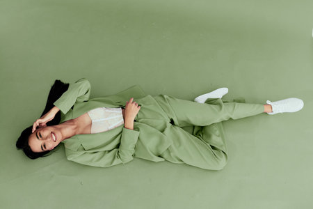 Young woman in green suit lying on floor smiling brightly, wearing white sneakers and lace top, casual stylish outfit with fresh and relaxed mood.の写真素材