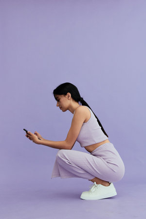 Young woman crouching and looking at smartphone, dressed in casual striped outfit and white...
