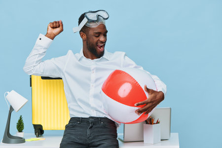 Happy young black man in white shirt enjoying fun with inflatable beach ball at office desk with suitcase and desk lamp on light blue background, cheerful mood, leisure conceptの写真素材