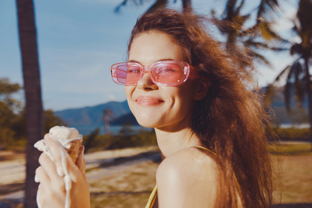 Cheerful young woman enjoying ice cream outdoors by the beach with palm trees in the background Bright sunny day enhances her playful moodの写真素材