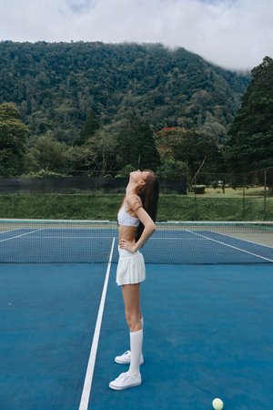 Young female tennis player stretching on a court, wearing a white sports outfit, conveying joy and confidence against a lush green mountain backdropの写真素材