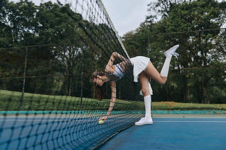 Stylish young woman in a tennis outfit, gracefully reaching for a ball near the net on a sunny day,...