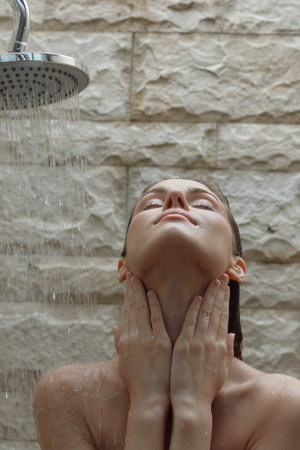 Woman enjoying relaxing shower with water falling on her face against textured stone wall,...