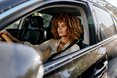 Young woman with curly hair driving car, wearing seatbelt and light jacket, focused and relaxed...