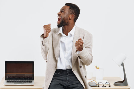 Enthusiastic smiling man in smart casual outfit celebrates success at his modern workspace with laptop and accessories on a white background, expressing happiness and motivation. Business lifestyle concept.の写真素材
