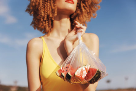 Woman holding a bag of fruit, standing against a clear blue sky in a vibrant yellow swimsuit