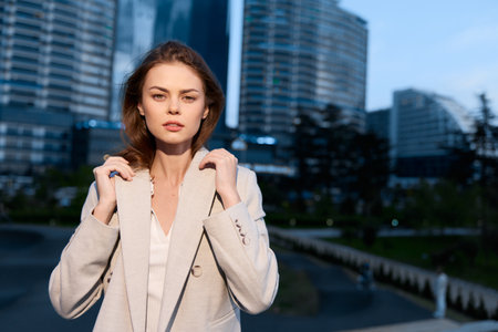 Young European woman in beige coat standing confidently in modern urban business district during daytime with office buildings background and clear skyの写真素材