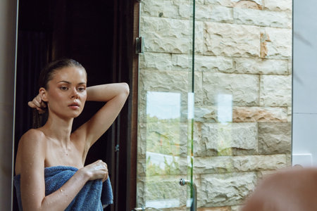 young woman wrapped in blue towel standing in bathroom with wet skin, natural light, modern interior, reflective glass surface, serene expressionの写真素材